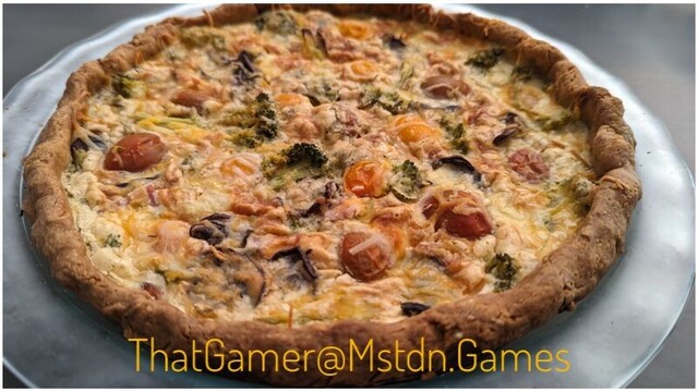 Photo shows a closeup of a homemade quiche on a serving plate. Visible on the quiche are sliced cherry tomatoes, mushrooms and grated cheese.
