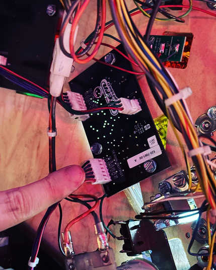 A printed circuit board under a pinball machine's playfield, with one connector being pointed out with a finger. 