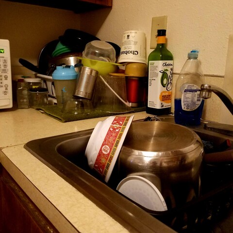 A plastic dish drainer on a kitchen counter tucked into a corner, a cloth drainer mat in front of that, and yet another drainer in the sink in front of that, all overfull with a chaotic assortment of cookware and cleaned recycling items, including a stainless steel dutch oven and the inner pot from an Instant Pot. 