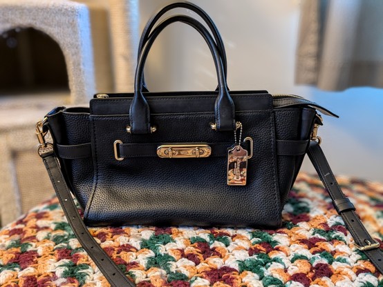 A Coach Swagger 27 black pebbled leather designer bag with gold hardware. Top handle and crossbody with a bold metal strap on the front and hangtag.