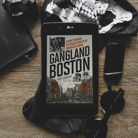 gangland boston by emily sweeney, a tour through the deadly streets of organized crime, ebook cover, portraying old streets of boston in the prohibition era. black-framed lg tablet is placed on a dark greyish wooden table, with various dark items strewn around, such as a crumped piee of balck paper, facemask, lighter, round shades.

