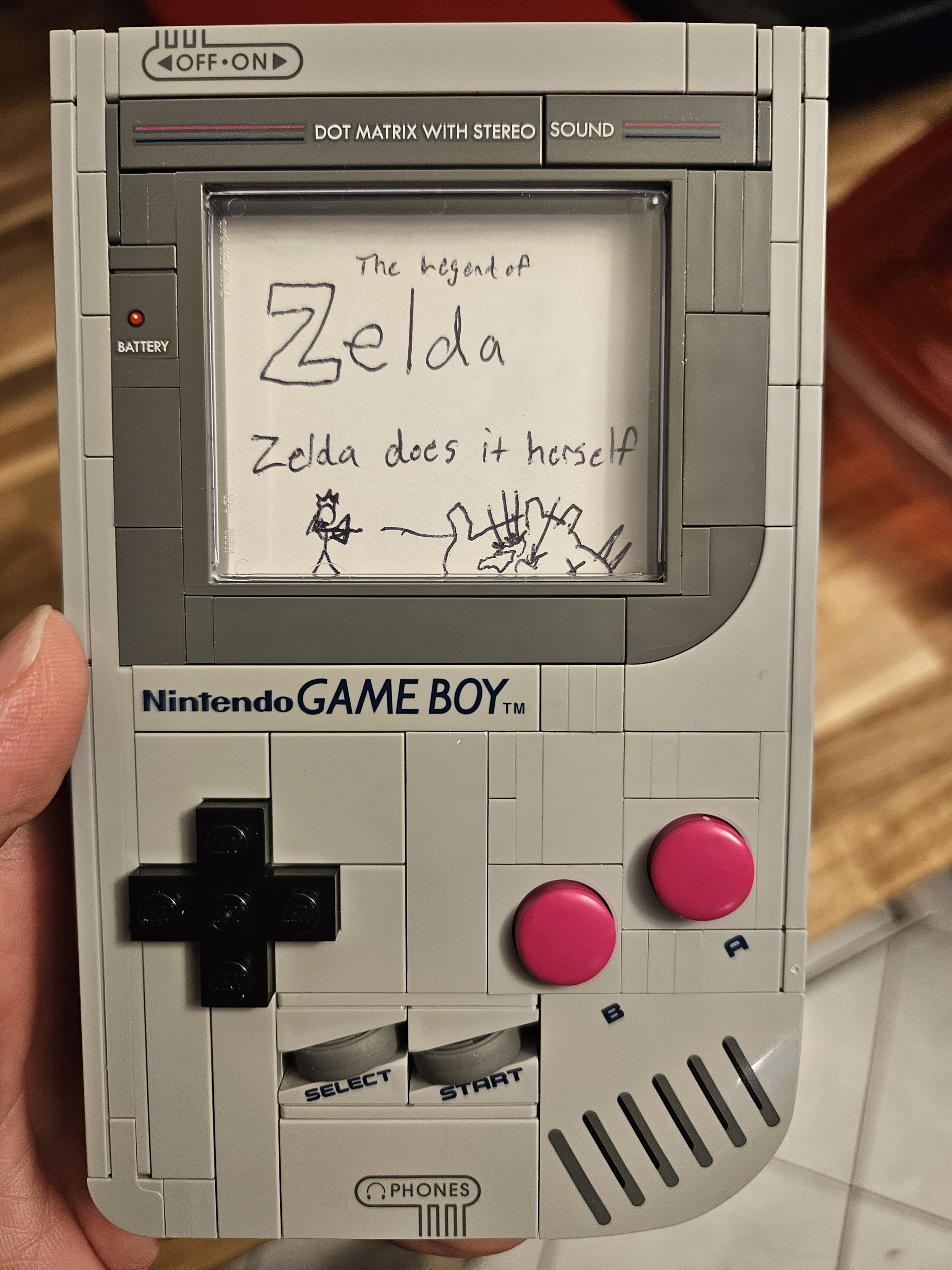 Photo of a Lego game boy with a piece of paper in the screen that reads "the legend of zelda: Zelda does it herseld"