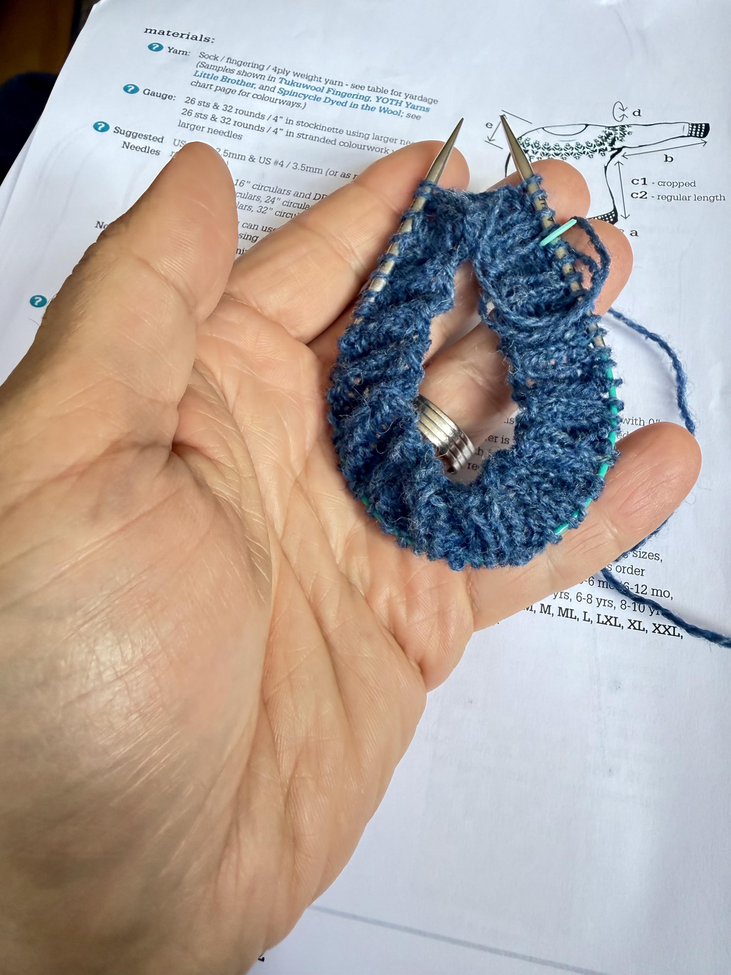 Huge, unbelievably sexy left hand holds a 9 inch circular needle with blue ribbing yarn on it. The hand has that soft-yet-rugged look that people just can’t resist. Yes.