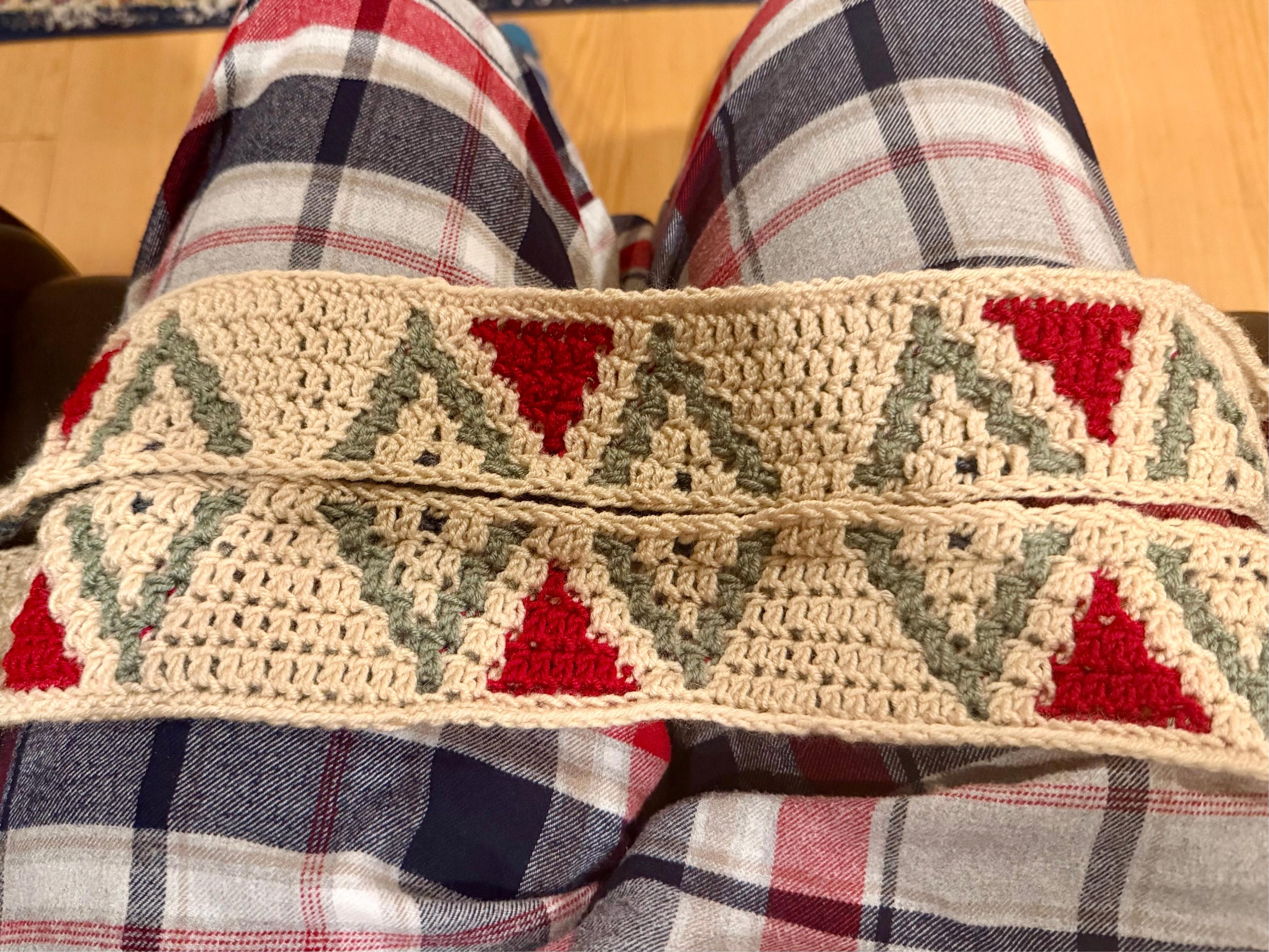 A close-up view of a crochet strip resting across someone’s lap clad in red, navy, and white plaid pajama trousers. The crochet work features a repeating geometric pattern of interlocking triangles — red pointing upward and sage green pointing downward — bordered by a beige background.