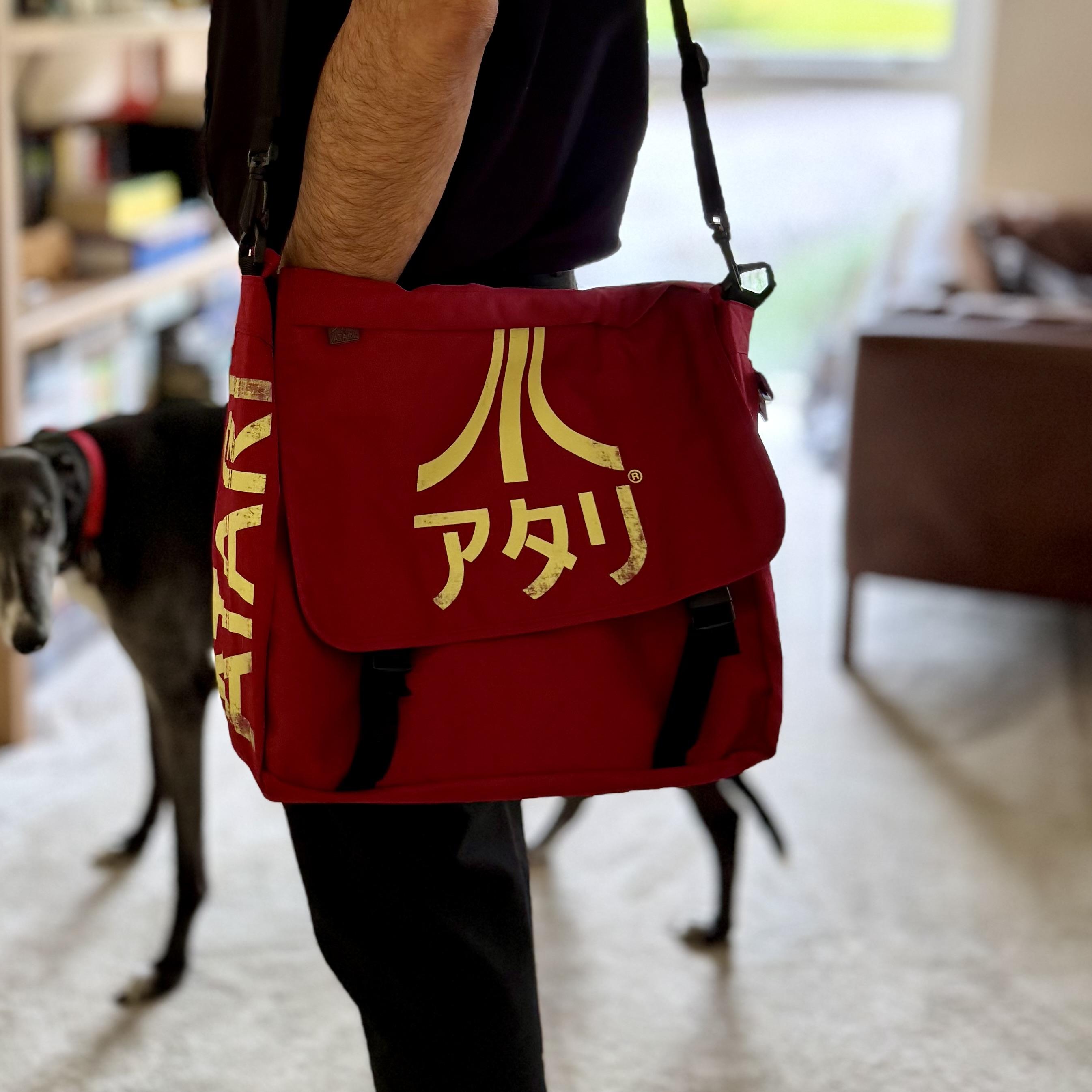 A debonair Arab man stands indoors wearing a red Atari shoulder bag with bold yellowish-gold lettering. The bag’s flap shows the classic Atari logo above Japanese katakana spelling “アタリ,” and the side panel features “ATARI” printed vertically. The bag hangs from a black adjustable strap, with black buckles securing the flap. In the softly lit background, a greyhound with a red collar walks across a pale carpet near a brown armchair and a bookshelf filled with assorted items.
