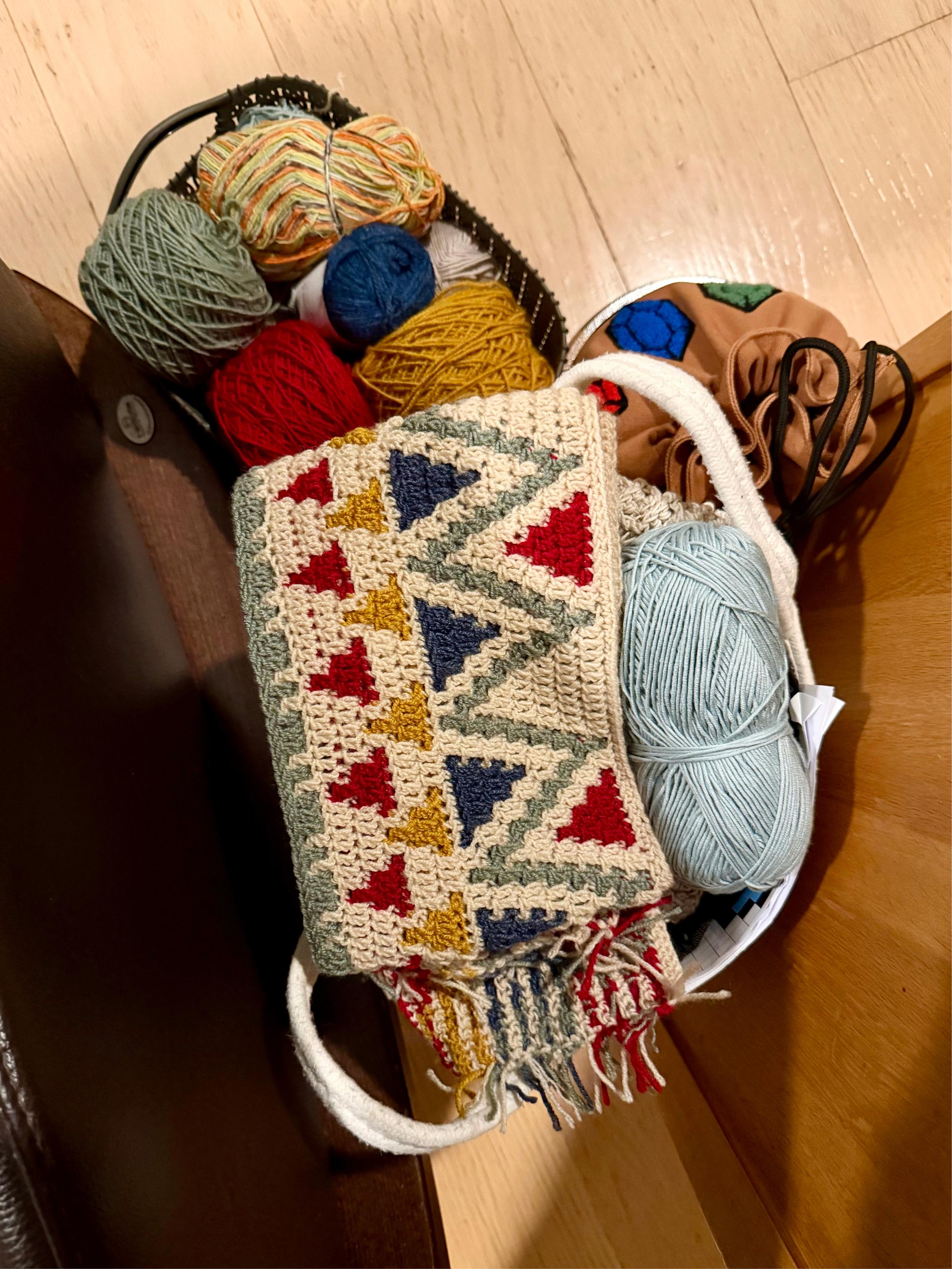 A basket filled with yarn balls and a partially completed crochet project. The yarns are in shades of green, red, yellow, blue, white, multicolored, and pale blue. The crochet piece draped over the basket features a geometric pattern of red, blue, and yellow triangles bordered by zigzag stripes in muted green, with fringes hanging from one end. The basket sits on a light wooden floor beside a wooden chair, and a tan fabric pouch with blue and green hexagonal patches is tucked alongside the yarns.