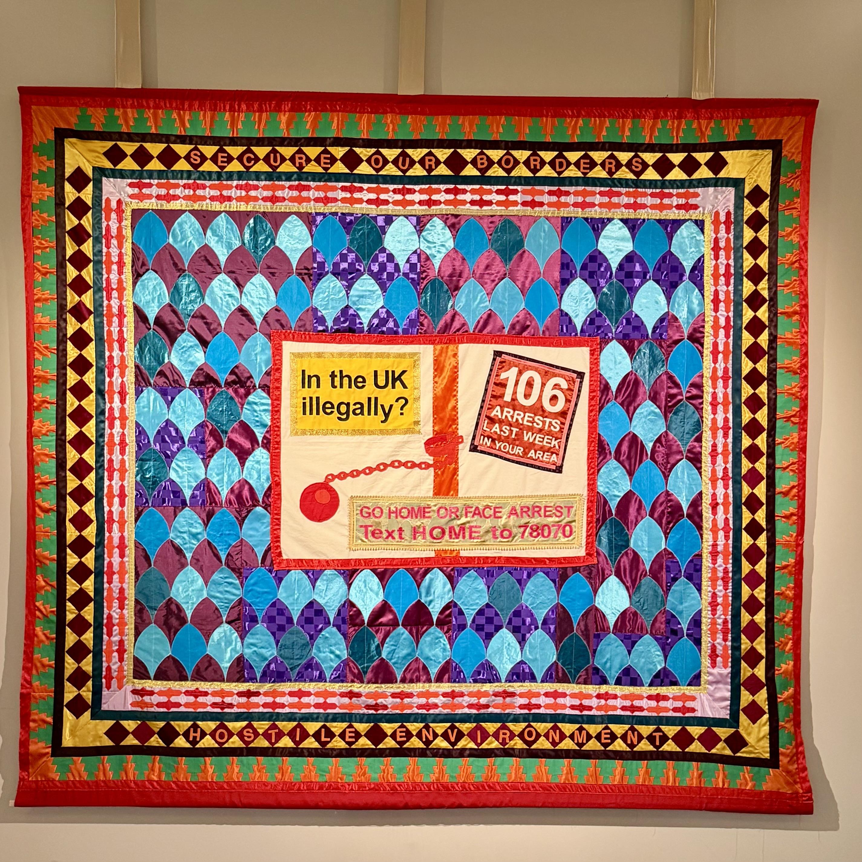 A large, colourful quilt artwork displayed on a wall. The quilt features a repeating teardrop pattern in shiny blue, purple, and turquoise fabrics bordered by geometric designs in red, yellow, and black. At its centre is a stitched replica of a UK Home Office “Go Home” campaign poster, reading “In the UK illegally? 106 arrests last week in your area. Go home or face arrest. Text HOME to 78070.” Around the edges are embroidered phrases “Secure Our Borders” and “Hostile Environment.” The piece contrasts the softness of textile craft with the harshness of its political message.