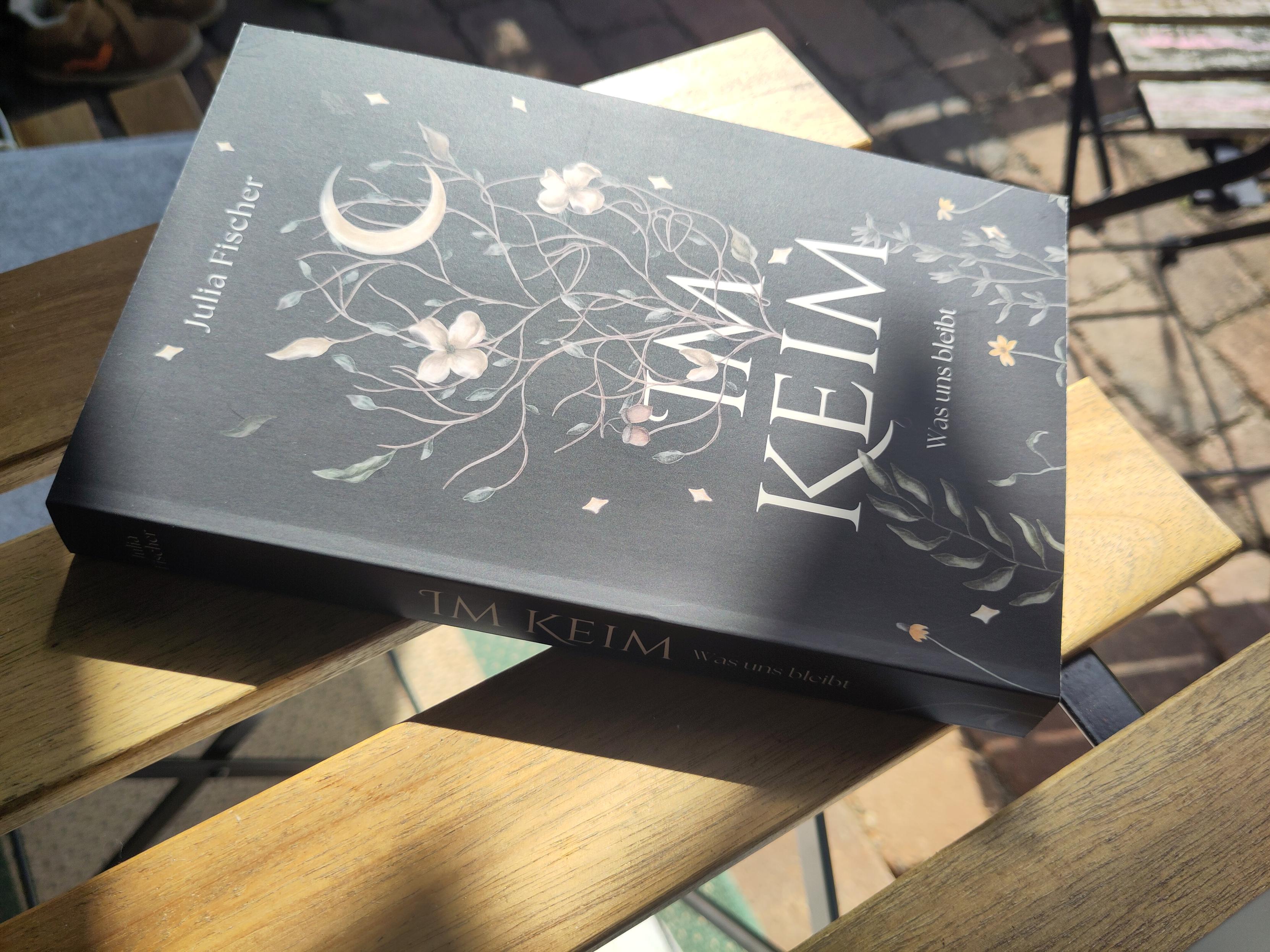 Das Buch "Im Keim - Was uns bleibt" von Julia Fischer liegt in der Sonne auf einem Holztisch.