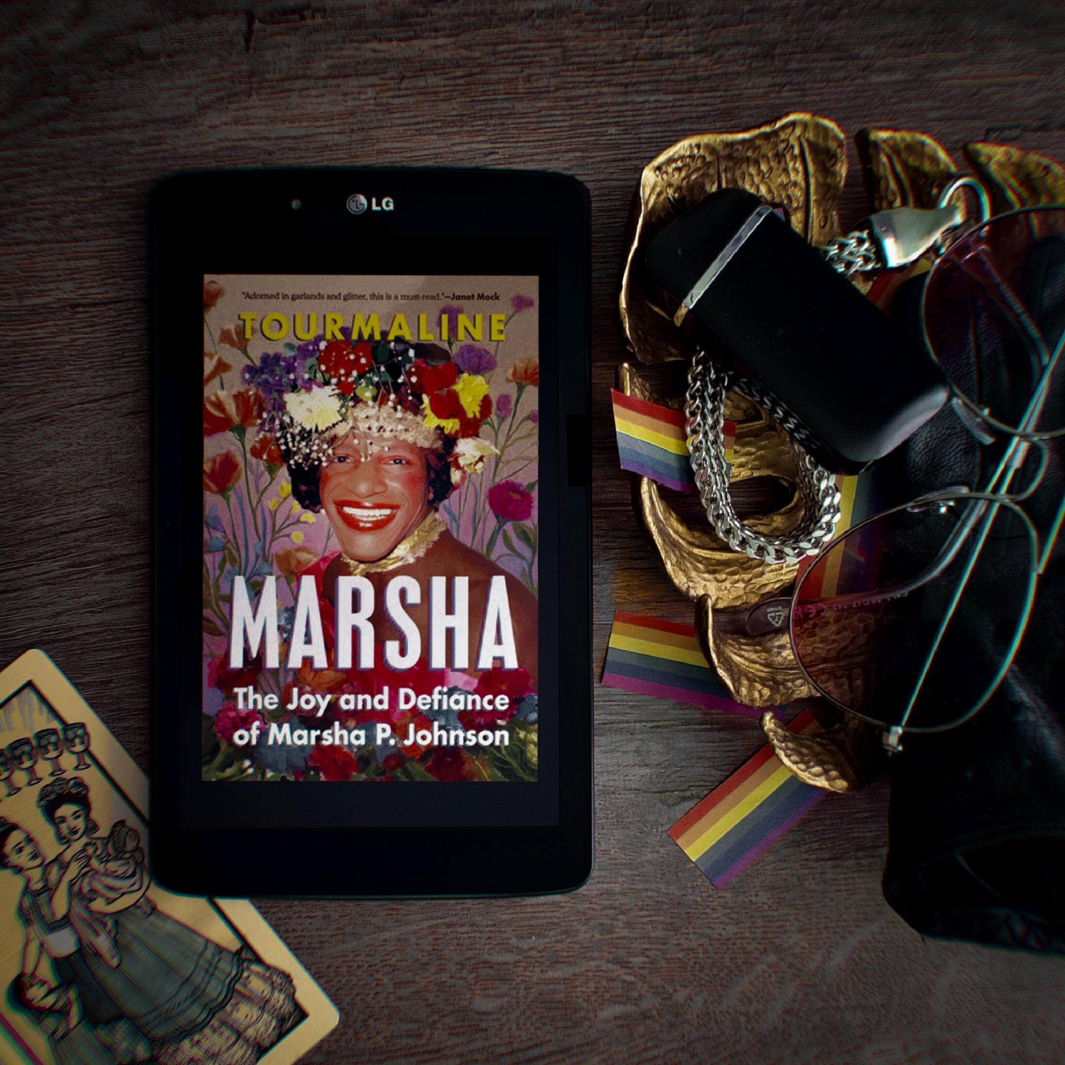 Marsha: the joy and defiance of marsha p. johnson by tourmaline, book cover, portraying marsha wearing a flower crown and floral dress, with floral background. she's smiling bright. black-framed lg tablet is placed on a dark wooden table, with various items around, such as monstera leaf shaped gold colored wooden bowl, some lgbtq flag confettis, lighter, shades, a tarot card. image and review by wannabekingpin aka vakaris, neither may be used for AI alteration or training.