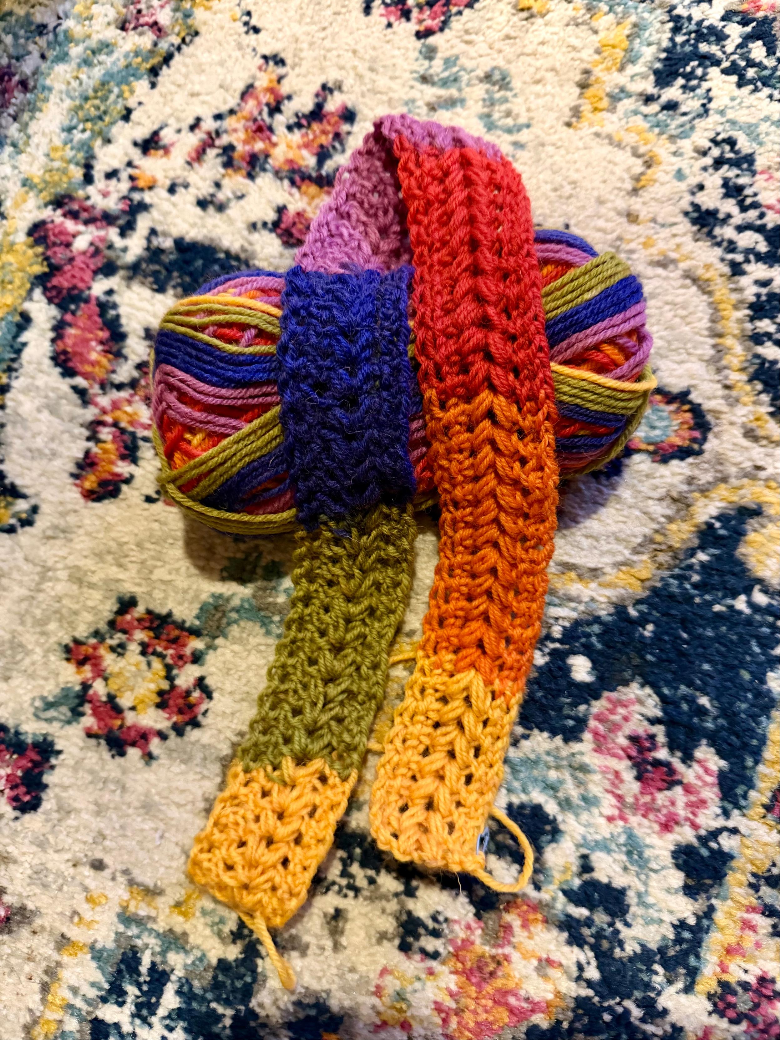 A ball of multicolored yarn rests on a patterned rug with floral motifs in cream, blue, pink, and yellow. Attached to the yarn is a crocheted strip showing a smooth gradient of colors — starting with pale yellow at one end, shifting through orange, olive green, deep blue, bright red, and finishing with soft purple at the other.