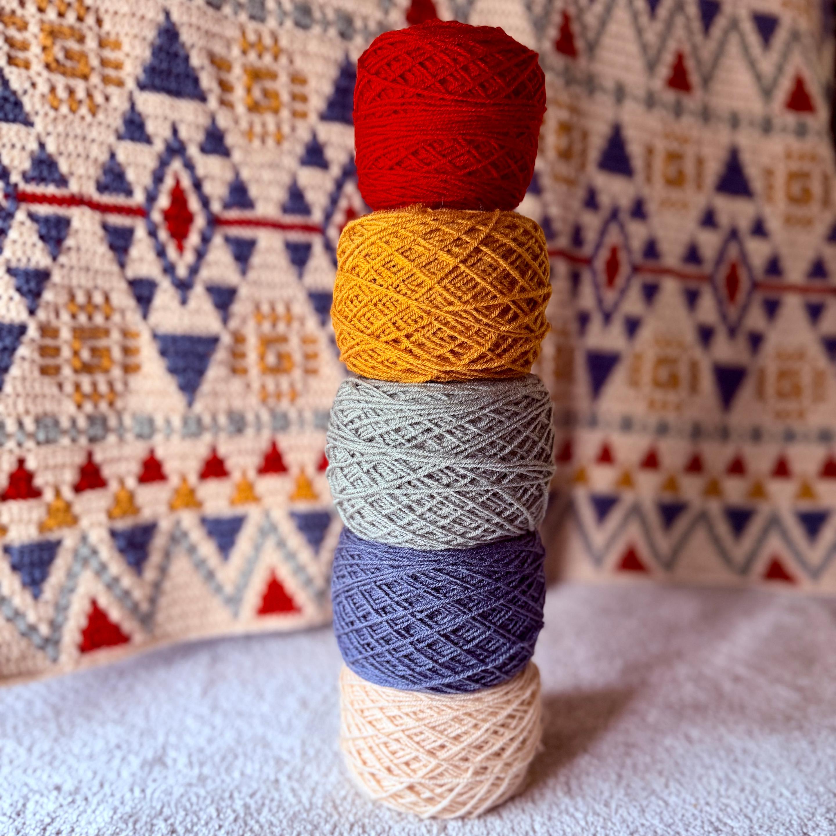 Stack of five yarn cakes in red, mustard, green, blue, and cream tones placed on a soft surface, with a geometric patterned crochet backdrop in coordinating colors.