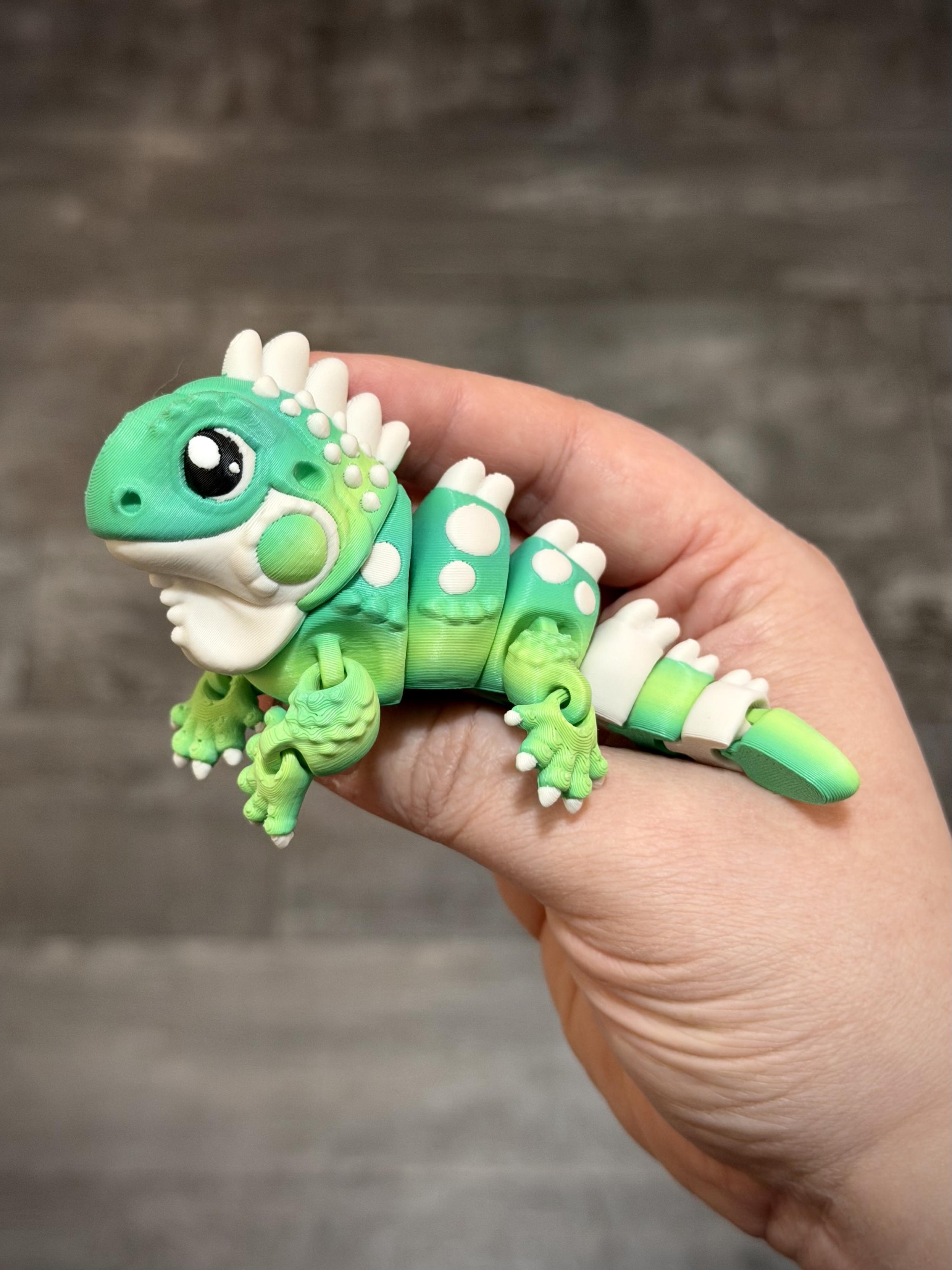 Small articulating flexi fidget toy of an iguana - green and blue green gradient body colour with bone white accents, held in the right hand on a neutral background m.