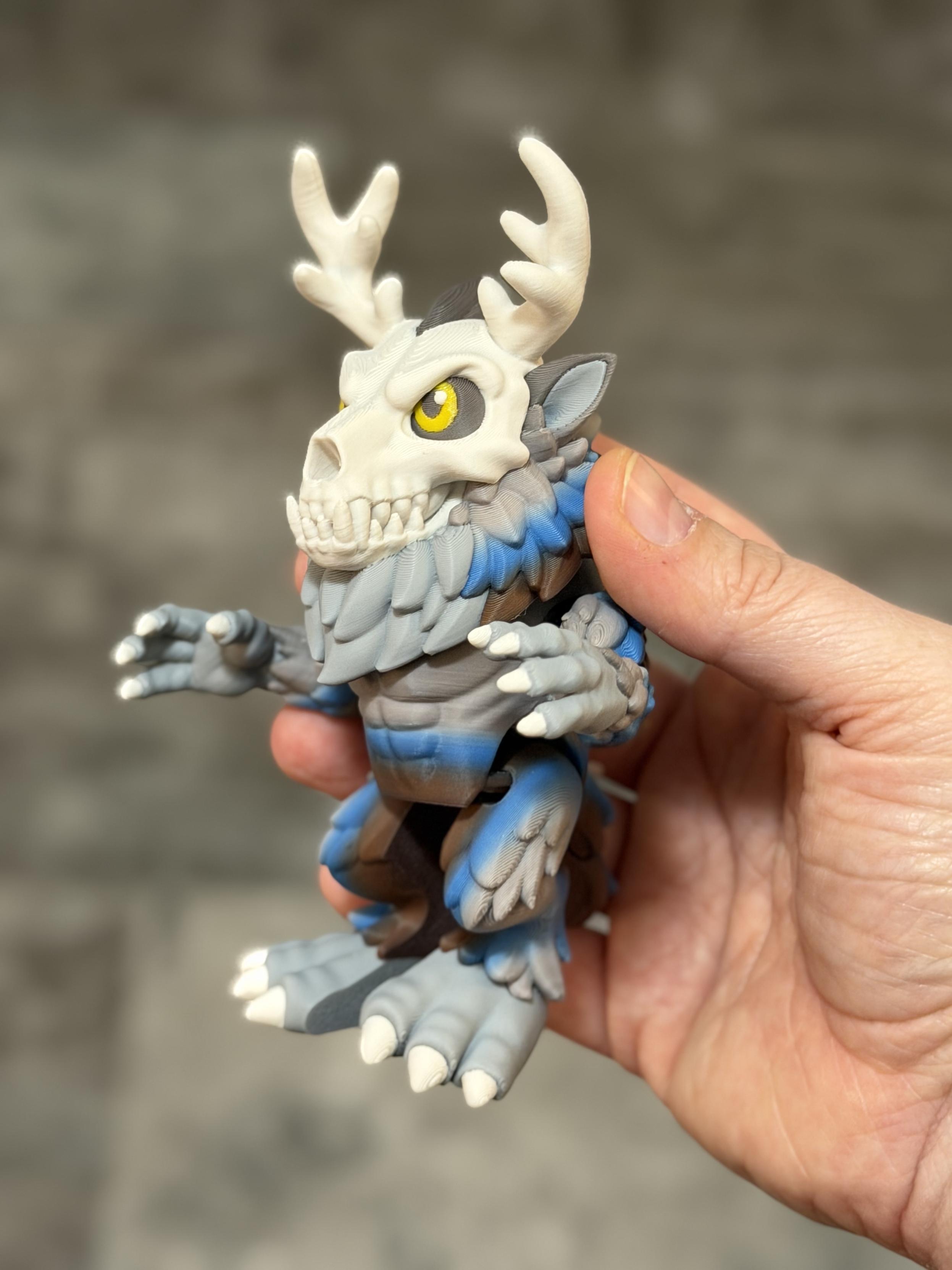 Hand holding a small figure of a wendigo, side profile. The eyes are yellow, with a bone white skull and blue grey and brown body.
