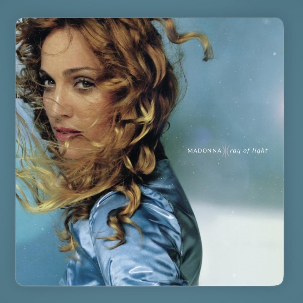 “Album cover for Ray of Light by Madonna. She’s shown in close-up, turning slightly with wind‑blown golden curls and a calm, confident expression. She wears a shiny blue jacket against a soft, ethereal blue background with subtle light specks.”