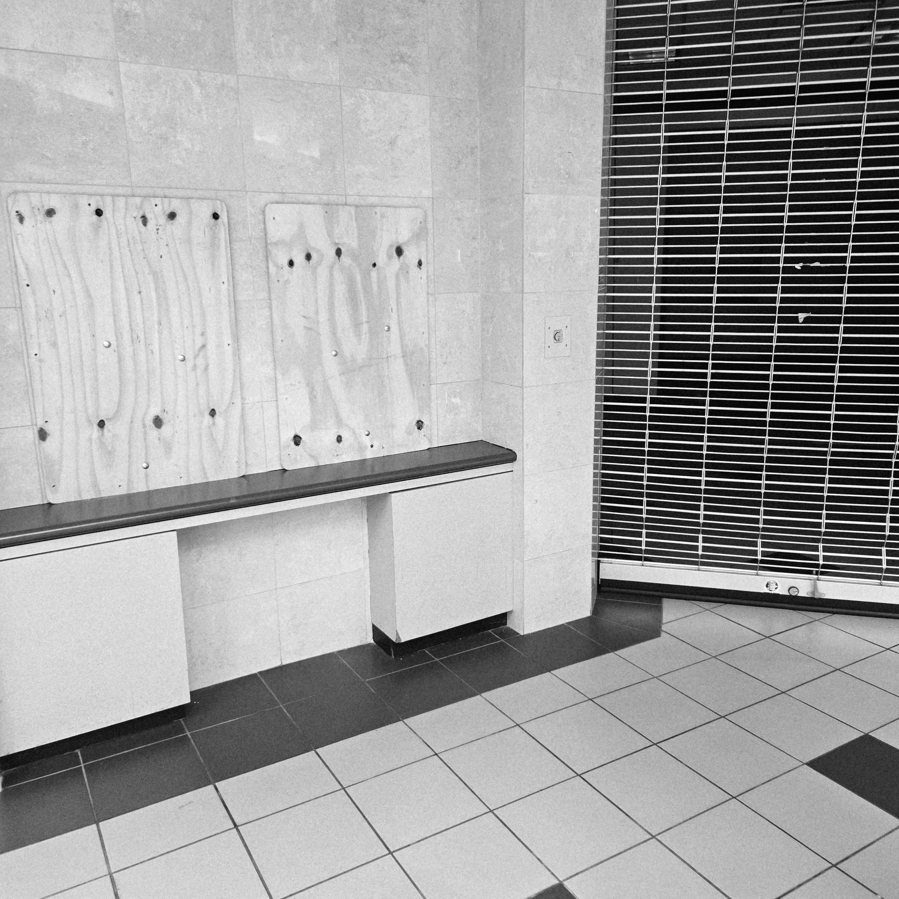 Black and white photograph of the corner between an abandoned, closed store with a garage door with slots blocking the entrance (right) to the storefront and a tiled wall (left). The tiled wall has two openings that are covered with wooden panels. This is above a shelf.