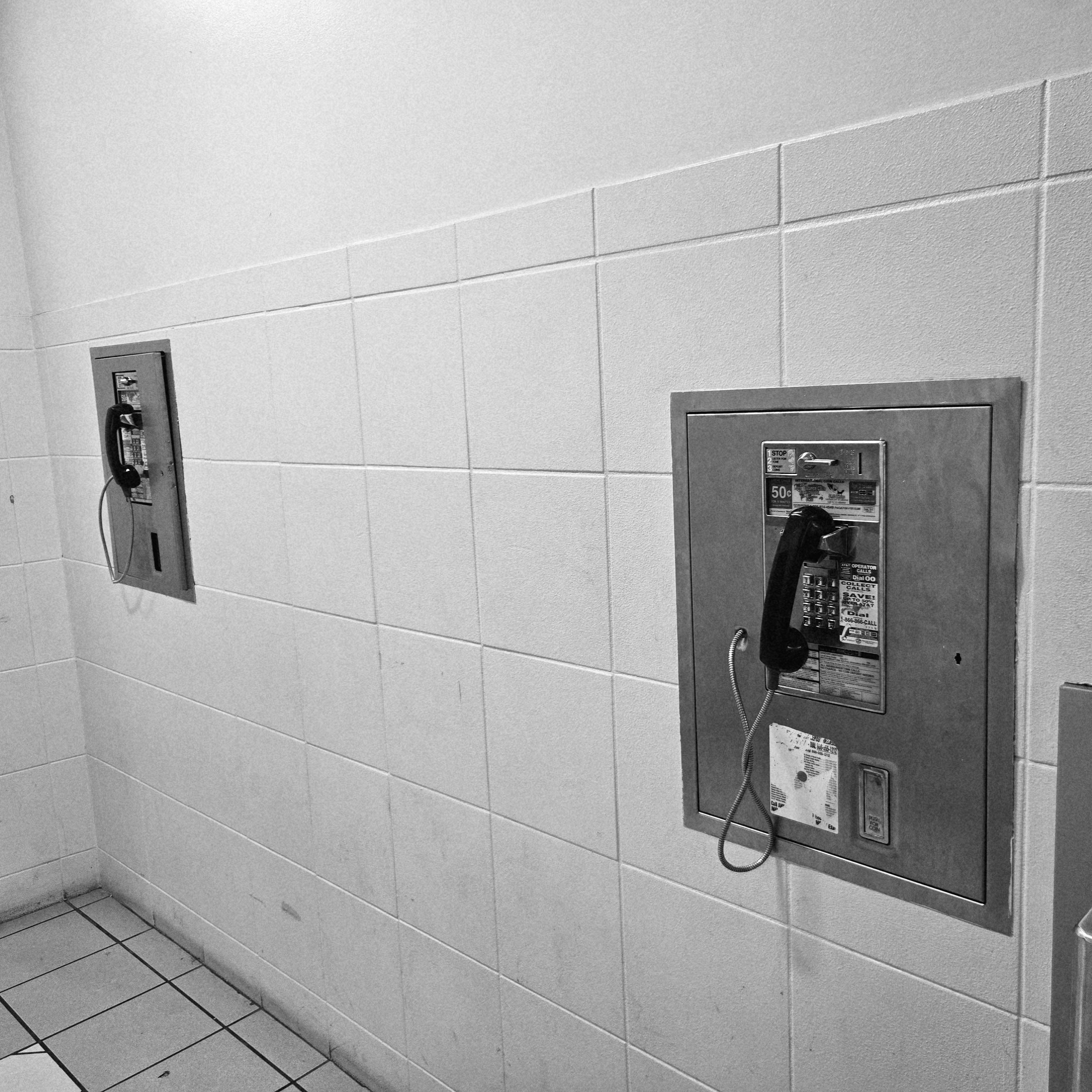 Black and white photograph of a tiled wall taken at an angle with two pay phones embedded into the wall separated by about 6 tiles.