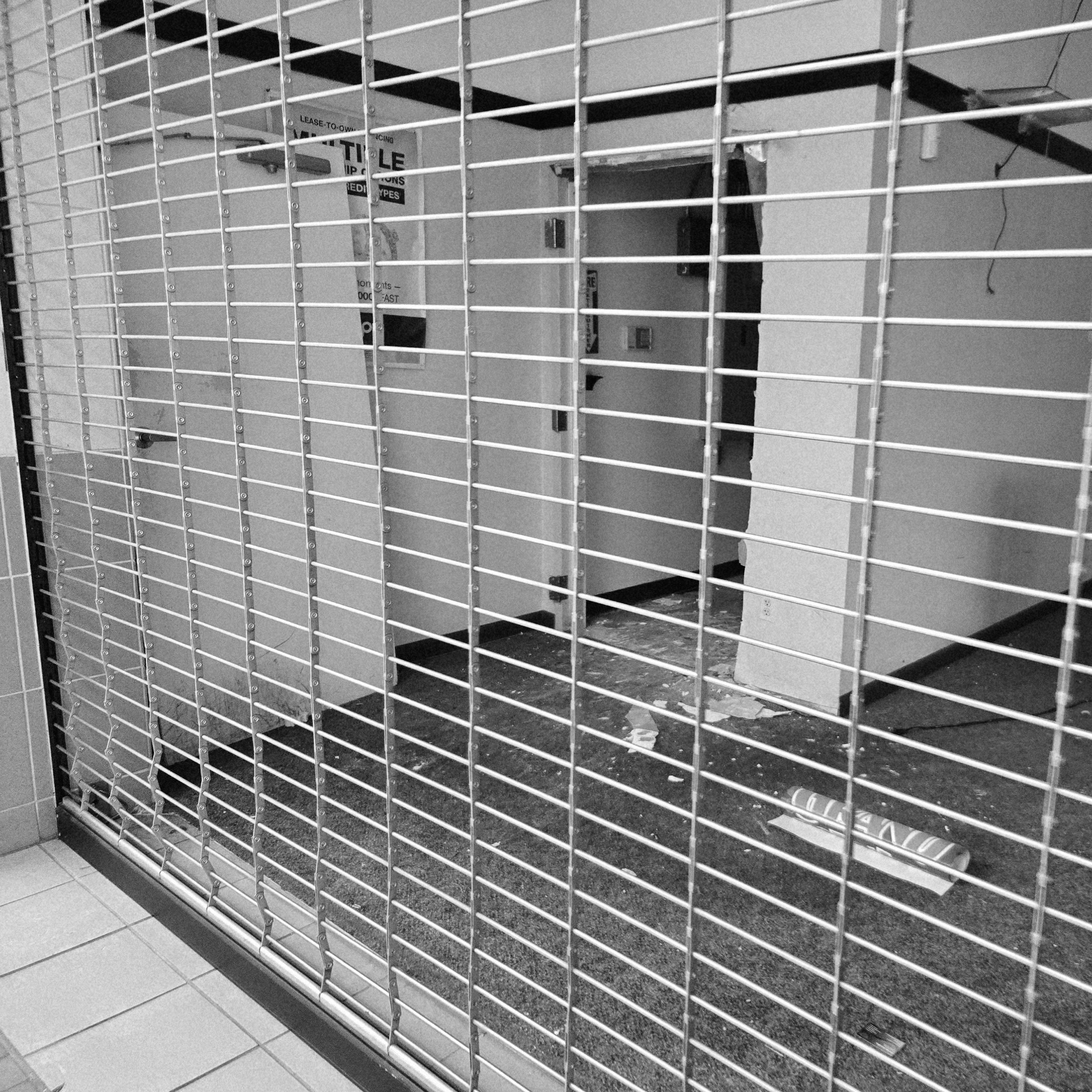 Black and white photograph of a storefront in a mall that has a garage door style cage brought down to block off the entrance. Inside of the storefront is trash on the ground, an open back room, and a partially obscured poster on the wall.
